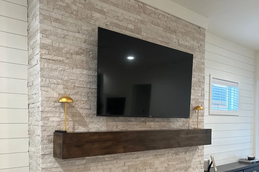 TV mounted on a stacked stone fireplace wall above a dark wood mantel and a linear electric fireplace